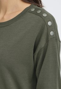 Gros plan sur un pull en jersey vert olive avec cinq boutons en argent le long de la couture de l'épaule porté par une personne.