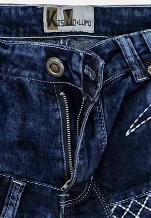 Jean en denim bleu foncé avec une texture déchirée, doté d'une fermeture éclair à l'avant et d'un bouton en métal. Étiquette de la marque visible à la taille.