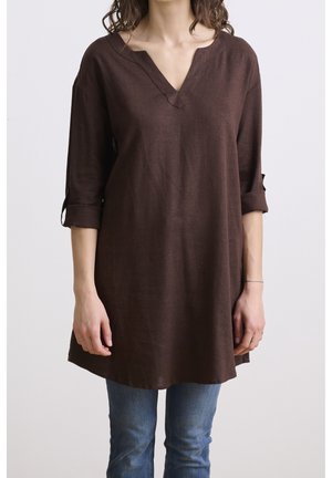 Femme portant une tunique ample à manches longues de couleur marron foncé avec les manches retroussées et un col en V, assortie à un jean bleu sur fond uni.