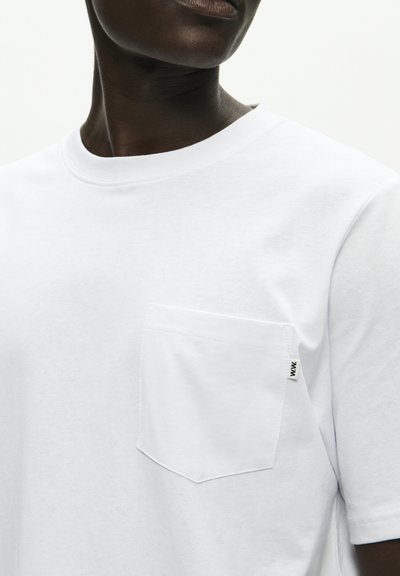 Nærbilde av person som har på seg en ensfarget hvit T-skjorte med brystlomme og et lite hvitt logomerke på lommens kant.