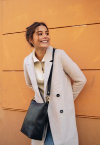 Helle beige, doppelt-knopfbare Jacke über einem cremefarbenen Reißverschluss-Pullover, kombiniert mit einer dunklen Leder-Schultertasche und blauen Jeans, vor einer orangen Wand.