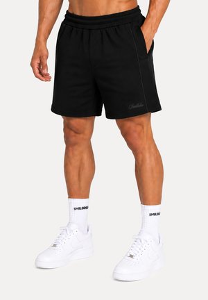 Uomo che indossa pantaloncini neri, calzini bianchi con testo nero e sneakers bianche, in piedi davanti a uno sfondo semplice.