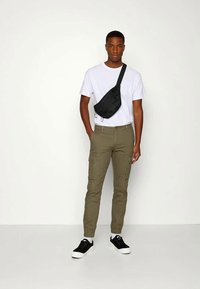 Άνδρας με λευκό t-shirt, ελαιοπράσινα cargo παντελόνια, μαύρη τσάντα crossbody, μαύρα αθλητικά παπούτσια και λευκές κάλτσες, που στέκεται μπροστά σε λευκό φόντο.