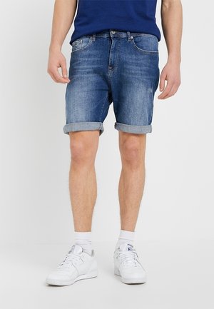 Homme portant un short en denim bleu retroussé, des chaussures de sport blanches, des chaussettes blanches et une chemise bleue, debout sur un fond blanc uni.