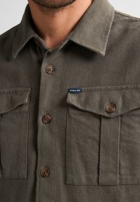 Dark green flannel shirt with a pointed collar, two chest pockets, brown buttons, and a small blue label on the left pocket.