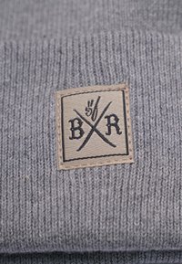 Harmaa neulottu pipo, jossa on teksturoitu pinta ja suorakulmainen vaaleanruskea patch, jossa on musta logo ja ristikkäiset viivat.