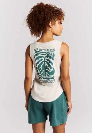 Jonge vrouw met krullend haar die een beige mouwloze top draagt met een groene zon- en bladprint en blauwe korte broek, staand in zijaanzicht.