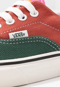 Niskoprofilowe sneakersy Vans mają górną część wykonaną z rdzawo-czerwonego sztruksu, ciemnozielone akcenty, białe oczka i teksturowaną gumową podeszwę z czarnym wykończeniem.