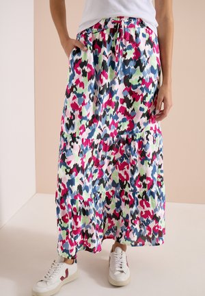 Femme portant une jupe longue fleurie colorée avec une taille élastique et des baskets blanches, debout avec une main dans la poche.