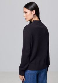 Schwarzer Strickpullover mit hohem Kragen und gerippten Bündchen, kombiniert mit blauen weit geschnittenen Jeans. Die Textur wirkt weich und glatt.
