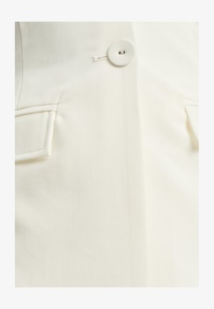 Tessuto color crema con due tasche cucite e chiusura con bottone. Texture liscia con una leggera lucentezza e design sartoriale.