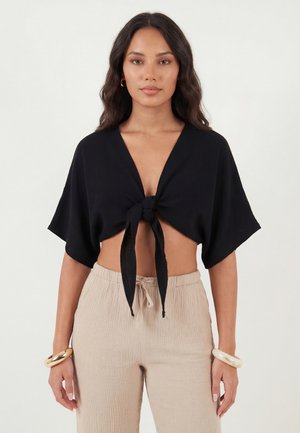 Žena s dlhými tmavými vlasmi, ktorá má oblečený čierny krátky crop top s krátkym rukávom, zviazaný vpredu a béžové nohavice s vysokým pásom na šnúrku, s náramkami.