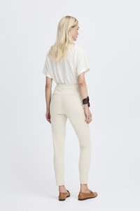 Béžové slim-fit kalhoty s hladkou texturou, se zadními kapsami. V kombinaci s světlou krátkým rukávem, texturovanou blůzou a hnědými botami.