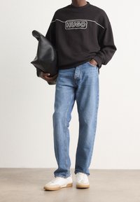 Sudadera negra con el logo "HUGO", jeans azules de pernera recta y zapatillas blancas con suelas de goma, sosteniendo un bolso negro de gran tamaño.