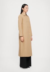 Beige Doppelreiher-Mantel mit taillierter Passform, spitzem Kragen, langen Ärmeln und seitlichen Taschen. Getragen über schwarzen weiten Hosen.