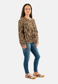 Donna in piedi con indosso una giacca a portafoglio con stampa leopardata, jeans skinny blu e sneakers beige con strisce bordeaux su sfondo bianco.