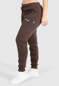 Detailaufnahme einer Person in braunen Jogginghosen mit Reißverschlusstaschen und weißen Sneakern, die vor einem schlichten hellen Hintergrund steht.