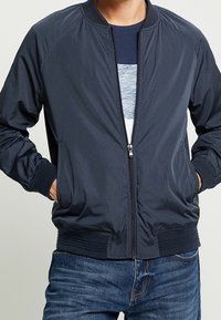 Marine Bomberjacke mit Reißverschluss, gerippten Ärmeln und Bund; zeichnet sich durch eine glatte Textur und lässiges Design aus. Getragen über einem blauen Hemd.