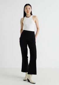 Top sem mangas branco combinado com calças pretas de cintura alta e boca de sino. A modelo usa botas de tornozelo de salto em off-white, exibindo um design minimalista.