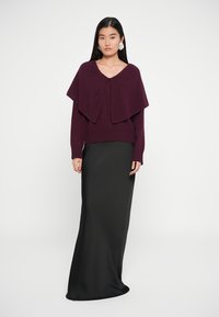 Maglione a maglia bordeaux con un design a strati su una gonna nera, lunga e dalla texture liscia. L'outfit è abbinato a orecchini oversize.