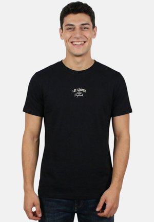 Homme souriant debout face à l’avant, portant un t-shirt noir Lee Cooper Originals et un jean foncé sur un fond uni clair.