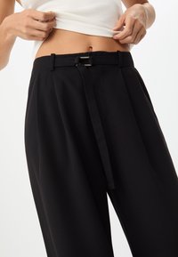 Pantaloni neri a gamba larga con una texture liscia, dotati di vita annodata e passanti per cintura. Design minimalista, adatti per un abbigliamento casual o semi-formale.