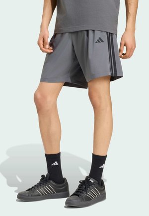 Person, der bærer grå sportsshorts med sorte striber og logo, sorte sokker og sorte Adidas-sneakers, står mod en lys baggrund.