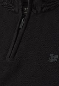 Gros plan d’un pull en maille noire avec une fermeture éclair au quart et un petit logo carré sur la poitrine gauche.