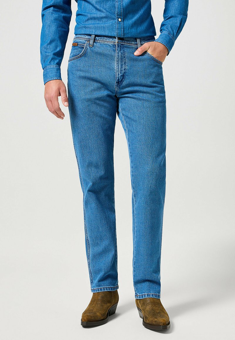 Jeans de mezclilla azul con un corte recto, que presentan un diseño estándar de cinco bolsillos y una superficie texturizada, llevados con botas marrones.