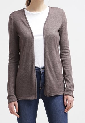 Femme portant un cardigan marron ouvert sur un t-shirt blanc et un jean bleu, debout devant un fond clair uni.