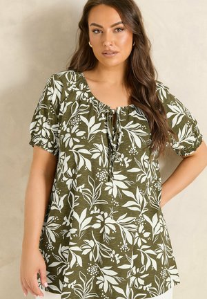 Vrouw met lang bruin haar die een olijfgroene tuniek met witte bloemenprint en korte pofmouwen draagt, staand tegen een beige achtergrond.