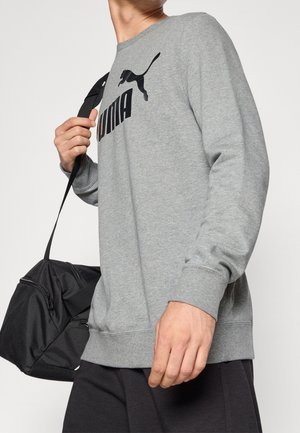 Person iført en grå Puma-sweatshirt og sorte bukser, bærende en sort duffel bag over skulderen mod en ensfarvet baggrund.