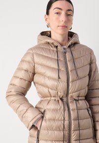 Giacca a vento beige con texture a righe verticali, zip frontale, cappuccio con coulisse e vita regolata. Presenta due tasche laterali e accessori in contrasto.