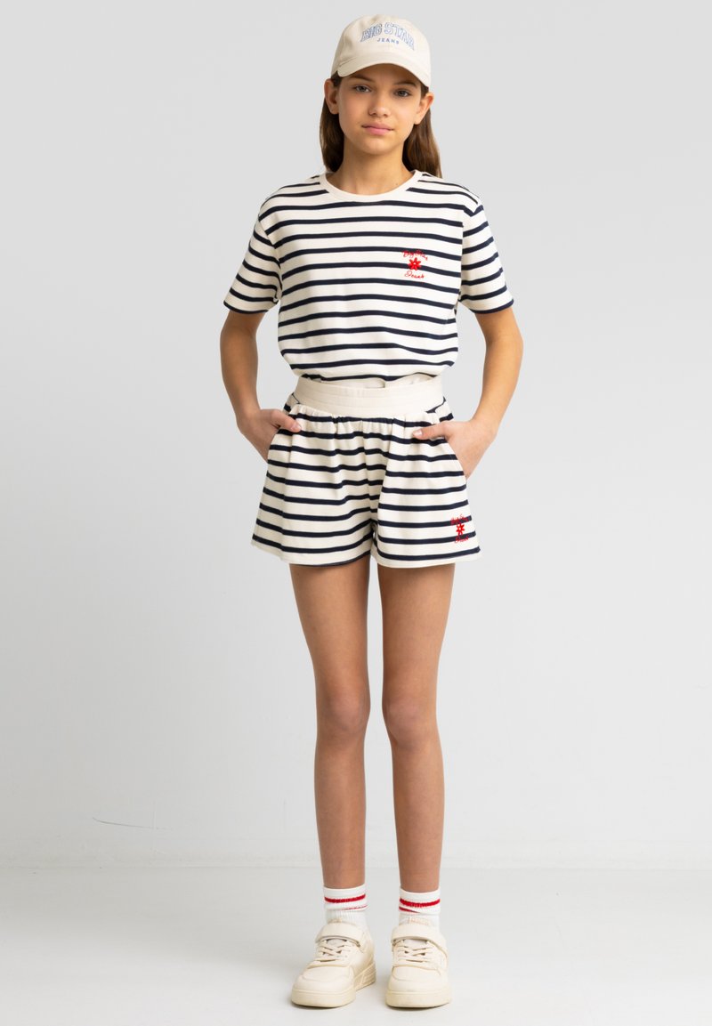 Pasiasta bawełniana koszulka i szorty w granatowym i kremowym kolorze z haftowanym czerwonym wzorem, połączone z beżową czapką i kremowymi trampkami.