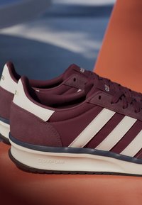 Paire de baskets Adidas bordeaux avec des bandes blanches, semelle Cloudfoam et détails en suède au talon, posées sur une surface orange.