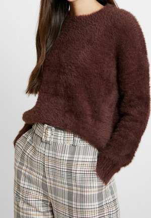 Femme portant un pull doux et pelucheux marron foncé et un pantalon à carreaux blanc taille haute avec une ceinture assortie, mains dans les poches.