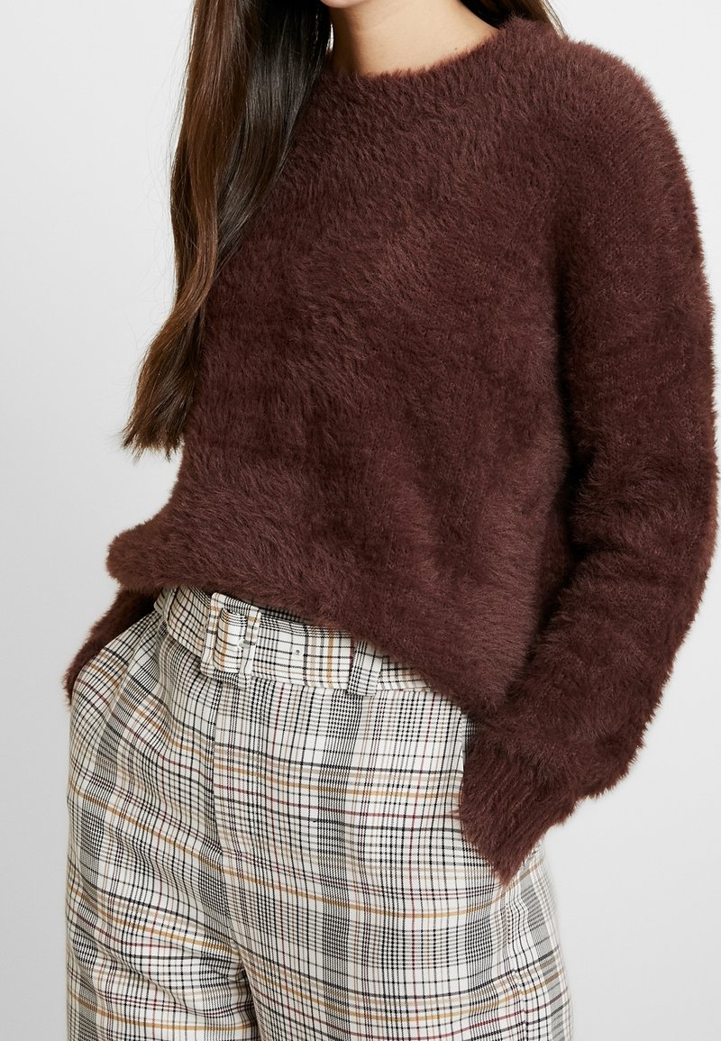 Femme portant un pull doux et pelucheux marron foncé et un pantalon à carreaux blanc taille haute avec une ceinture assortie, mains dans les poches.