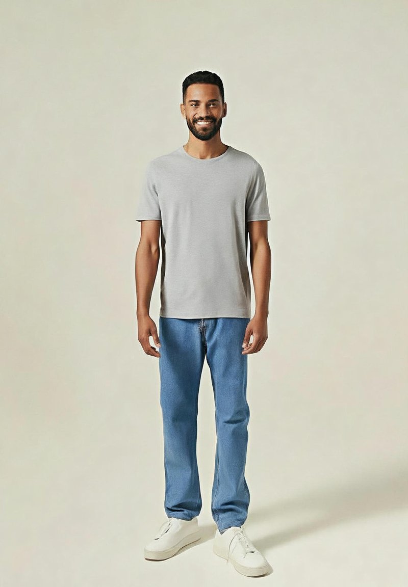 Mężczyzna uśmiechający się, stojący w jasnoszarej koszulce, niebieskich dżinsach i białych trampkach na jednolitym beżowym tle.