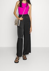 Vrouw die een felroze mouwloze top draagt, zwarte satijnen broek met wijde pijpen, zwarte sandalen met bandjes en een kleine beige crossbodytas draagt.