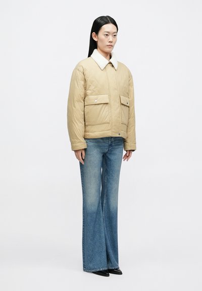 Beežilt pehmendusega jope valge kraega, kahe esitasku ja klambritega, kantud kulunud lameda siniste teksade ja mustade kingadega.