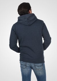 Dunkelblaues Kapuzenpullover mit großer, weicher Kapuze und geripptem Saum. Der Stoff wirkt strukturiert und ist kombiniert mit hellblauen Jeans.