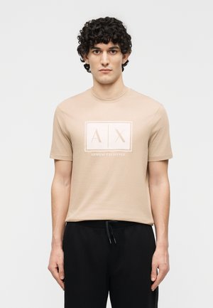 Άνδρας με σγουρά μαλλιά, που φοράει μπεζ μπλουζάκι Armani Exchange και μαύρο παντελόνι, στέκεται μπροστά σε ένα απλό φωτεινό φόντο.