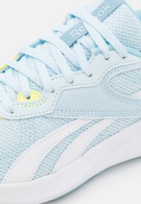 Světle modrá sportovní bota s texturovaným síťovaným svrškem, bílými akcenty, polstrovaným límcem a plochými tkaničkami s živým žlutým detailem.