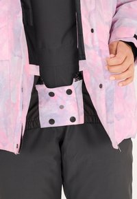 Bunte Jacke mit einem pinken und lila Aquarellmuster, die mit schwarzen Knöpfen und Reißverschlussverschlüssen über einer schwarzen Basisschicht befestigt ist.