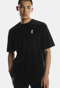 Fekete póló puha anyagból, fehér logóval a bal mellen. Rövid ujjú, laza szabású, minták vagy díszítések nélkül.