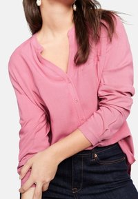 Camicia a maniche lunghe, rosa, con bottoni e scollatura a V, leggermente texturizzata. Indossata sopra jeans scuri, con una vestibilità rilassata e orlo curvo.