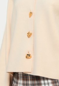 Απαλό ροζ πουλόβερ με τρία χρυσά, ακανόνιστου σχήματος κουμπιά στο πλάι. Μαλακή υφή, με τελείωμα που επικαλύπτει μια καρό φούστα.