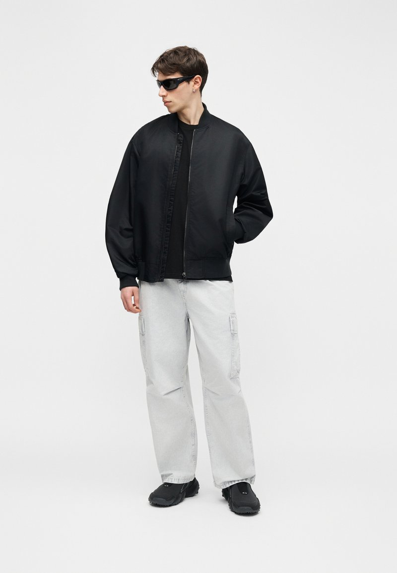 Junger Mann trägt schwarze Bomberjacke, schwarze Sonnenbrille, lockere hellgraue Cargohose und schwarze Schuhe, steht vor einem schlichten Hintergrund.