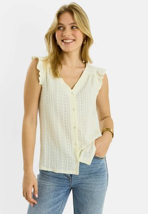 Mujer sonriente con cabello rubio lleva una blusa blanca sin mangas con ojales, botones y hombros con volantes, combinada con vaqueros azul claro y joyas de oro.