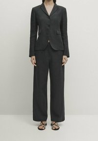 Woman wearing a dark gray tailored blazer with matching wide-leg pants and black strappy sandals, standing against a plain light background.